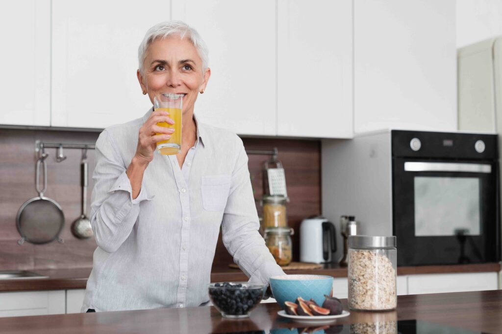 Fit Mature Woman Drinking Orange Juice With Vitamins While Having Breakfast At Home Kitchen. Middle Aged Housewife Eating Healthy Granola With Fruits And Oat Flakes
